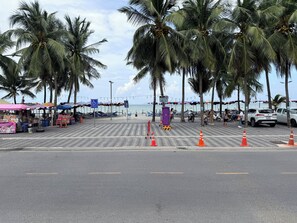 A beachside street with palm trees, beach umbrellas, and a pedestrian crossing. - Kelly Hotel Bangsaen (Mueang Chonburi)