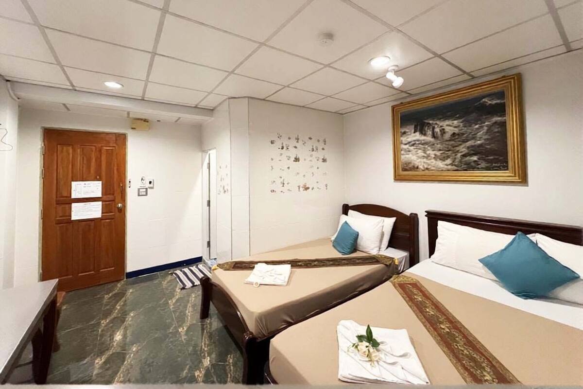 Habitación de hotel con dos camas individuales, una puerta de madera, pared blanca con un cuadro enmarcado y suelo de baldosas.