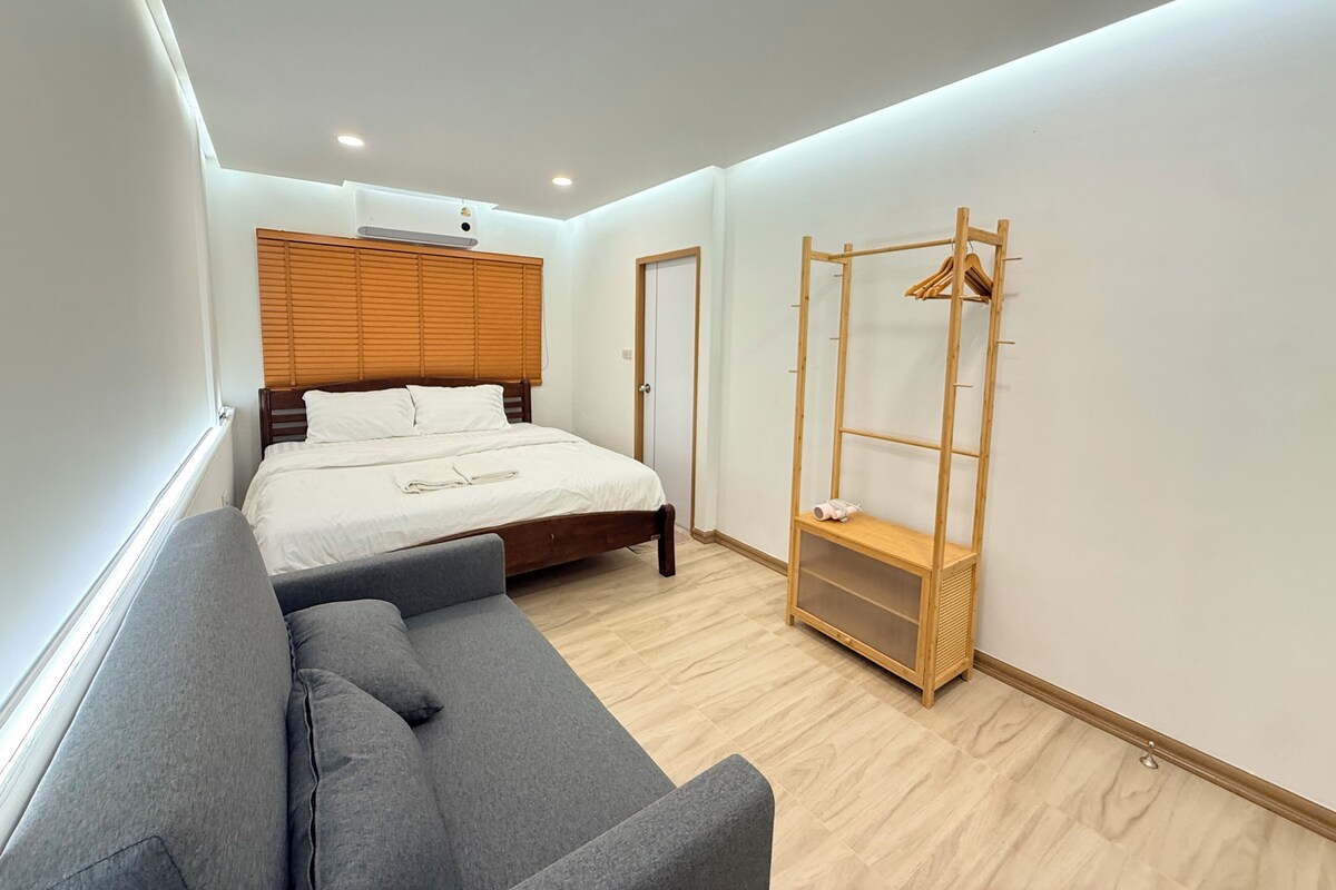 A bedroom with a bed, a grey sofa, a wooden wardrobe, and a wooden headboard.