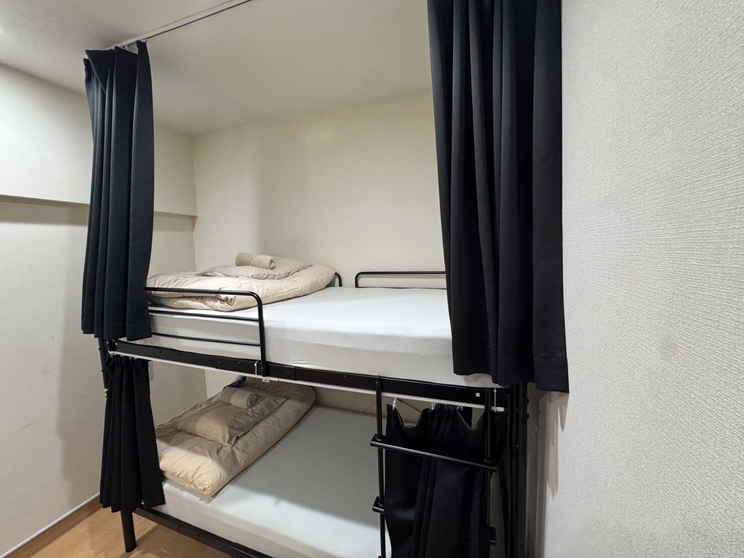 Una cama individual superpuesta con cortinas negras, un colchón blanco y una litera inferior con una manta color beige.