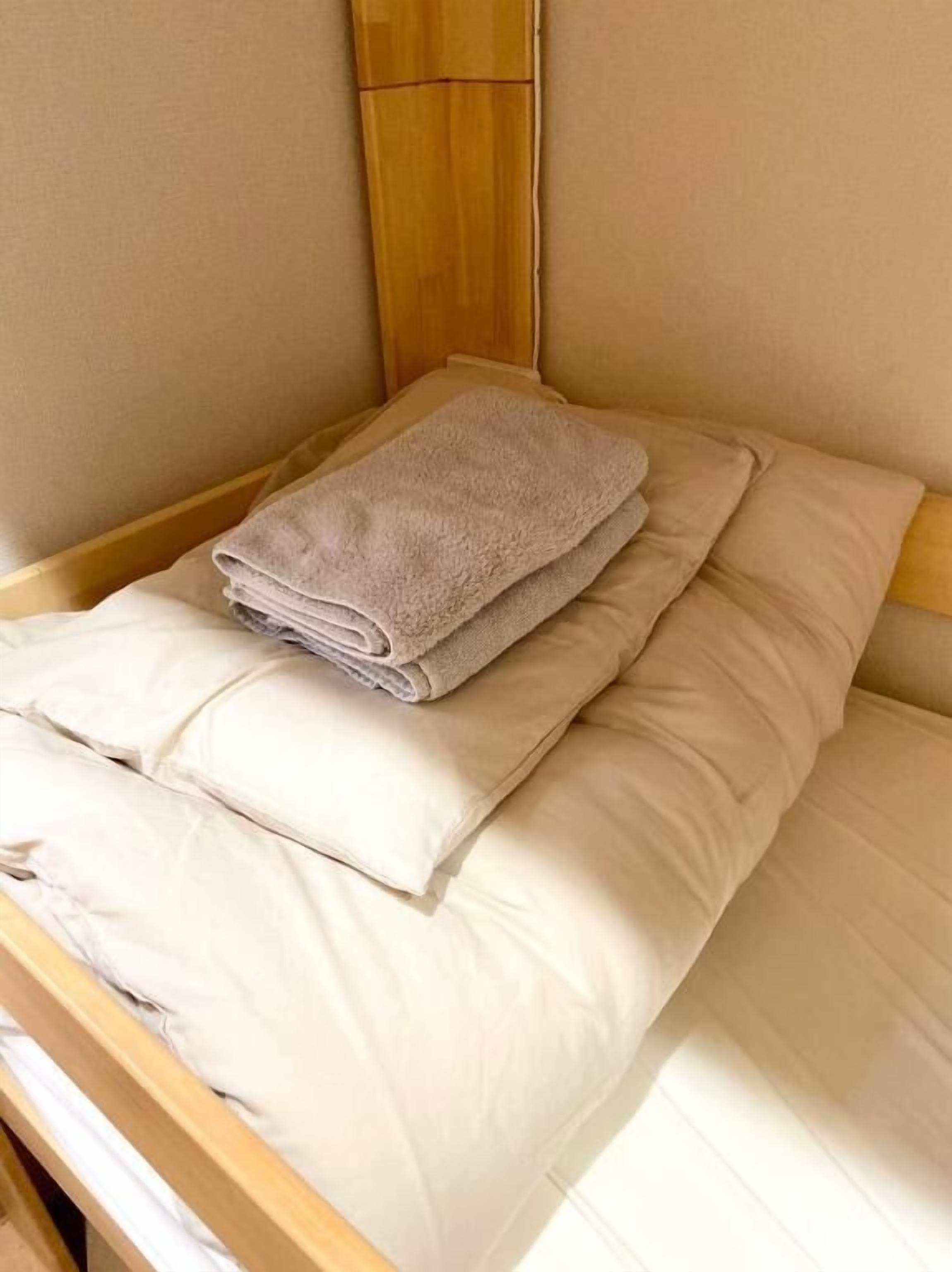 A bed with a folded towel and pillows, wooden headboard, and light-colored walls.