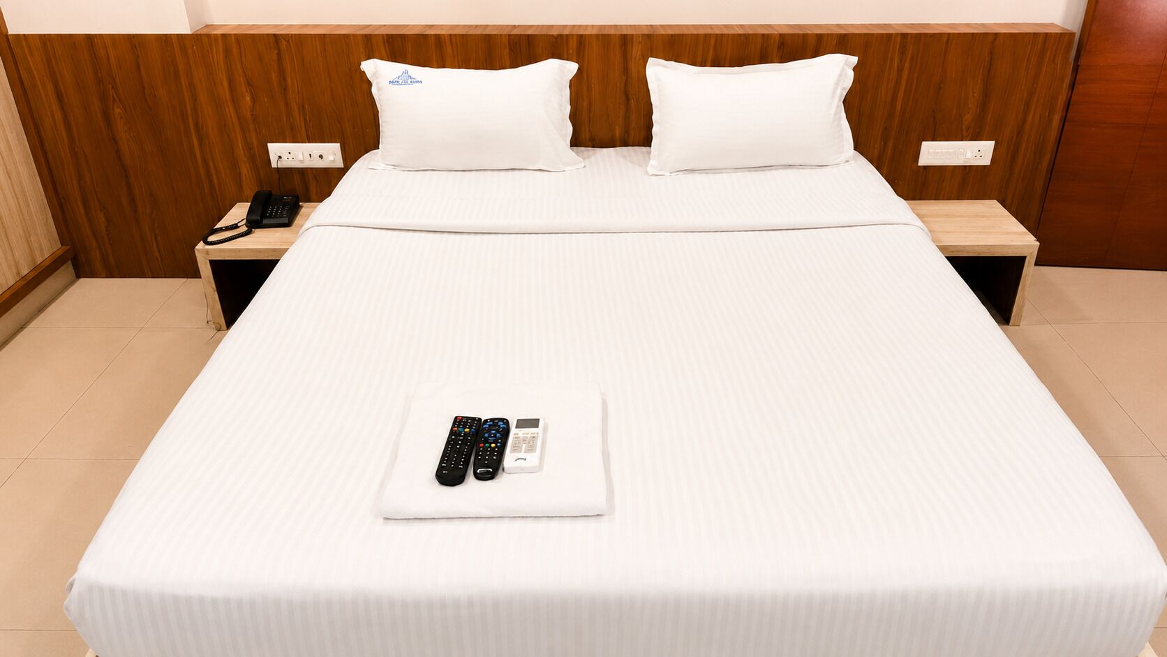 Habitación de hotel con una cama grande, dos almohadas, una mesita de noche con un teléfono y un control remoto sobre la cama.