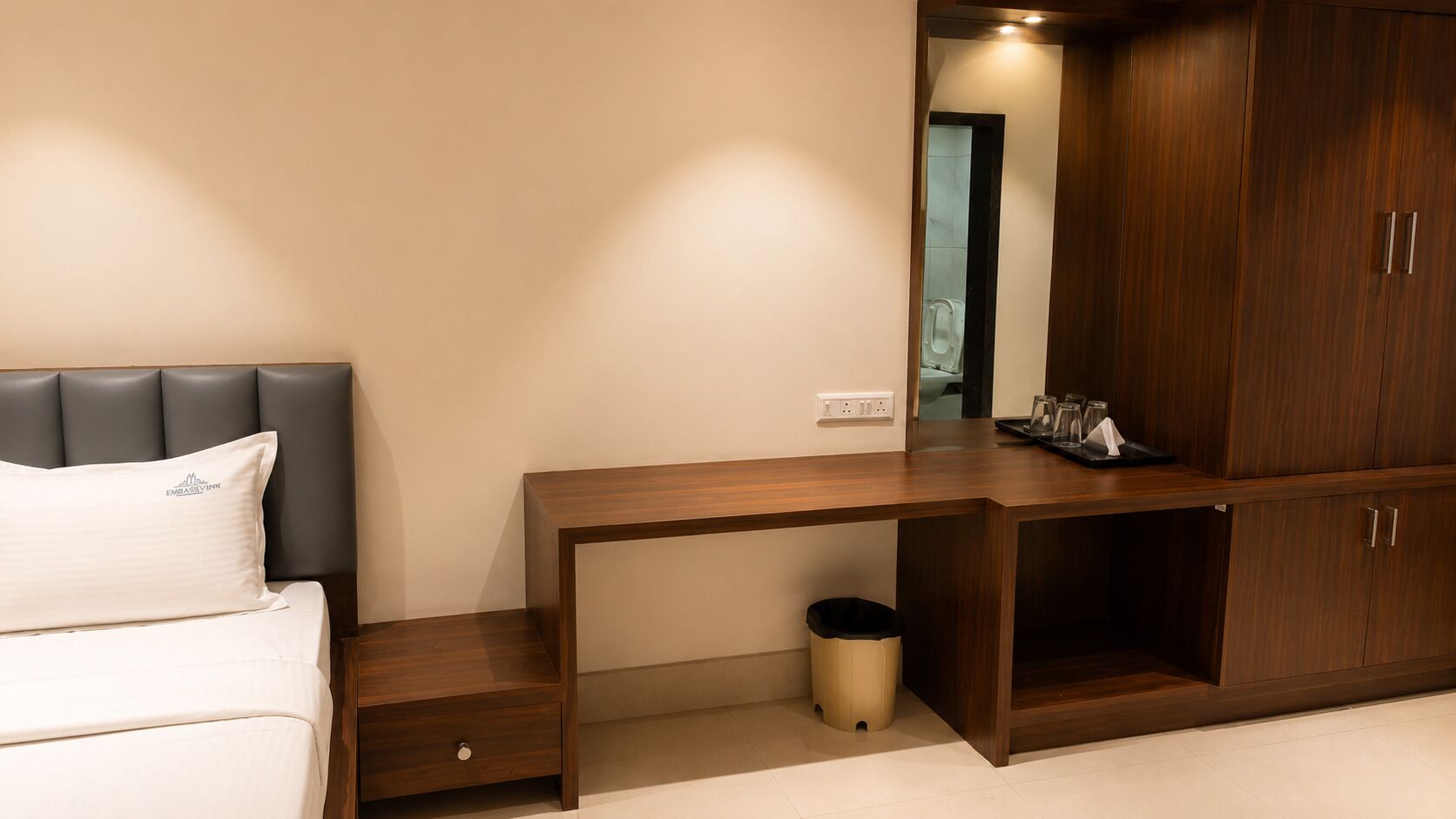 A bedroom with a bed, a wooden desk, a trash bin, and a wardrobe.