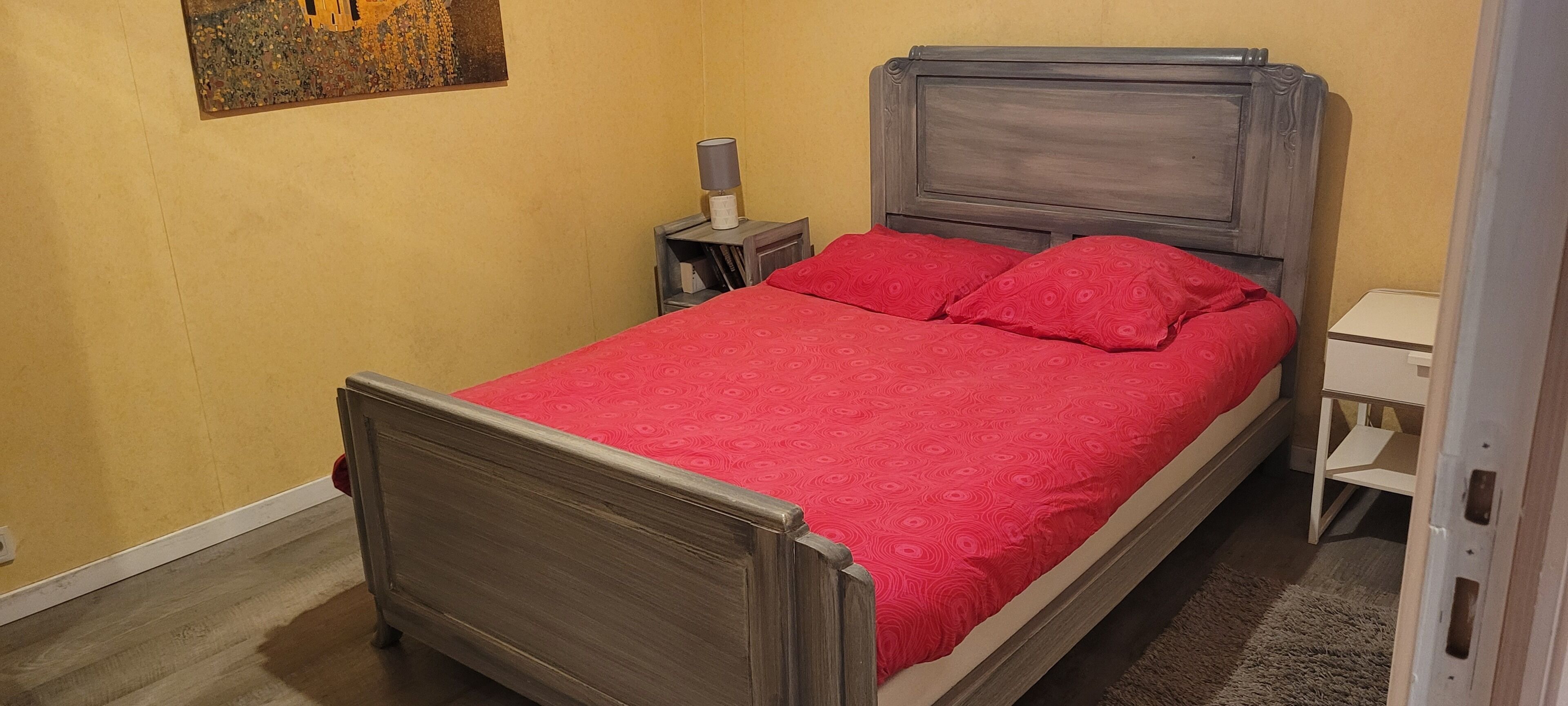 A single bed with a wooden frame and a red bedspread, a bedside table with a lamp, and a small white cabinet.