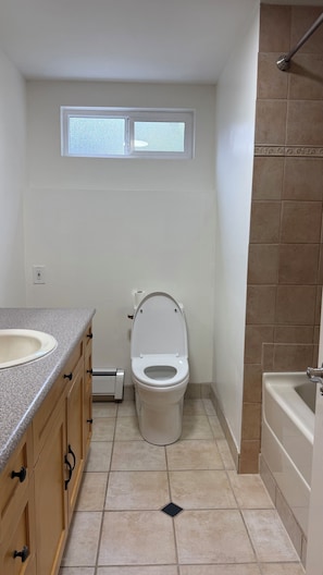Baño con inodoro, lavamanos y bañera. Hay una ventana sobre el inodoro y una zona de ducha a la derecha.