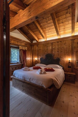 A wooden-paneled bedroom with a large bed, a window with curtains, and a nightstand. - Casa Chalet Cortina – Spacious Living and Dolomite Views (Cortina d´Ampezzo)