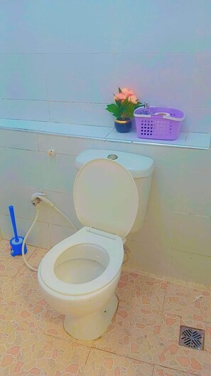 Eine Toilette mit Bidet-Ausstattung, ein blauer Reinigungsbürste, ein violetter Korb und ein Blumentopf in einem Badezimmer mit gefliesten Wänden und Boden.