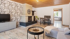 A living room with a stone wall, a fireplace, a television, and a round table with a clock. - Holiday Apartment An Der Lempe with Wi-Fi (Hofgeismar)