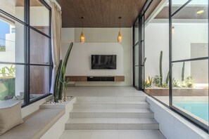 A modern interior with a staircase leading to a living area, a flat-screen TV mounted on the wall, and a pool visible through large windows. - Uluwatu 2 Bedrooms villa with Rooftop (Kec. Kuta Sel)