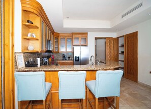 A kitchen with wooden cabinets, a granite countertop, a stainless steel refrigerator, and three bar stools. - Tessoro #1002 at Las Conchas – Elevated Luxury on the Sea of Cortez (Puerto Peñasco)