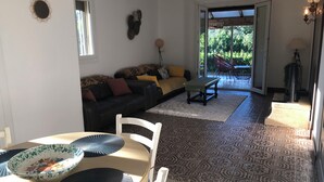 Ein Wohnzimmer mit einer Couch, einem Esstisch mit Stühlen, einem gemusterten Teppich und Blick durch eine Glastür auf einen Außenbereich.