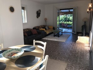Ein Wohnzimmer mit einer Couch, einem Esstisch mit Stühlen, einem gemusterten Teppich und Blick durch eine Glastür auf einen Außenbereich.