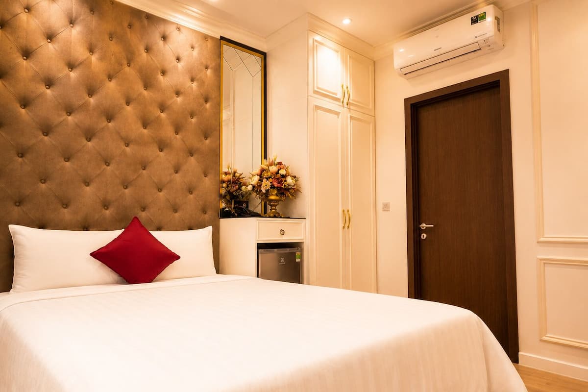 A hotel room with a tufted headboard, a bed with white bedding and a red pillow, a small bedside table with a vase of flowers, a wardrobe with a mirror, and a door with a wooden frame.