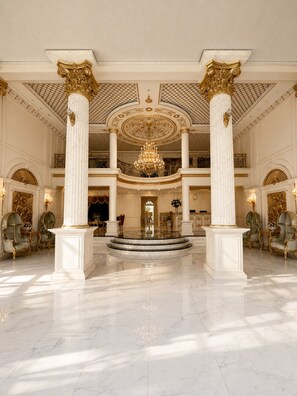 Un vaste hall d’entrée avec un sol en marbre, des colonnes richement décorées, un grand lustre et des éléments architecturaux classiques.