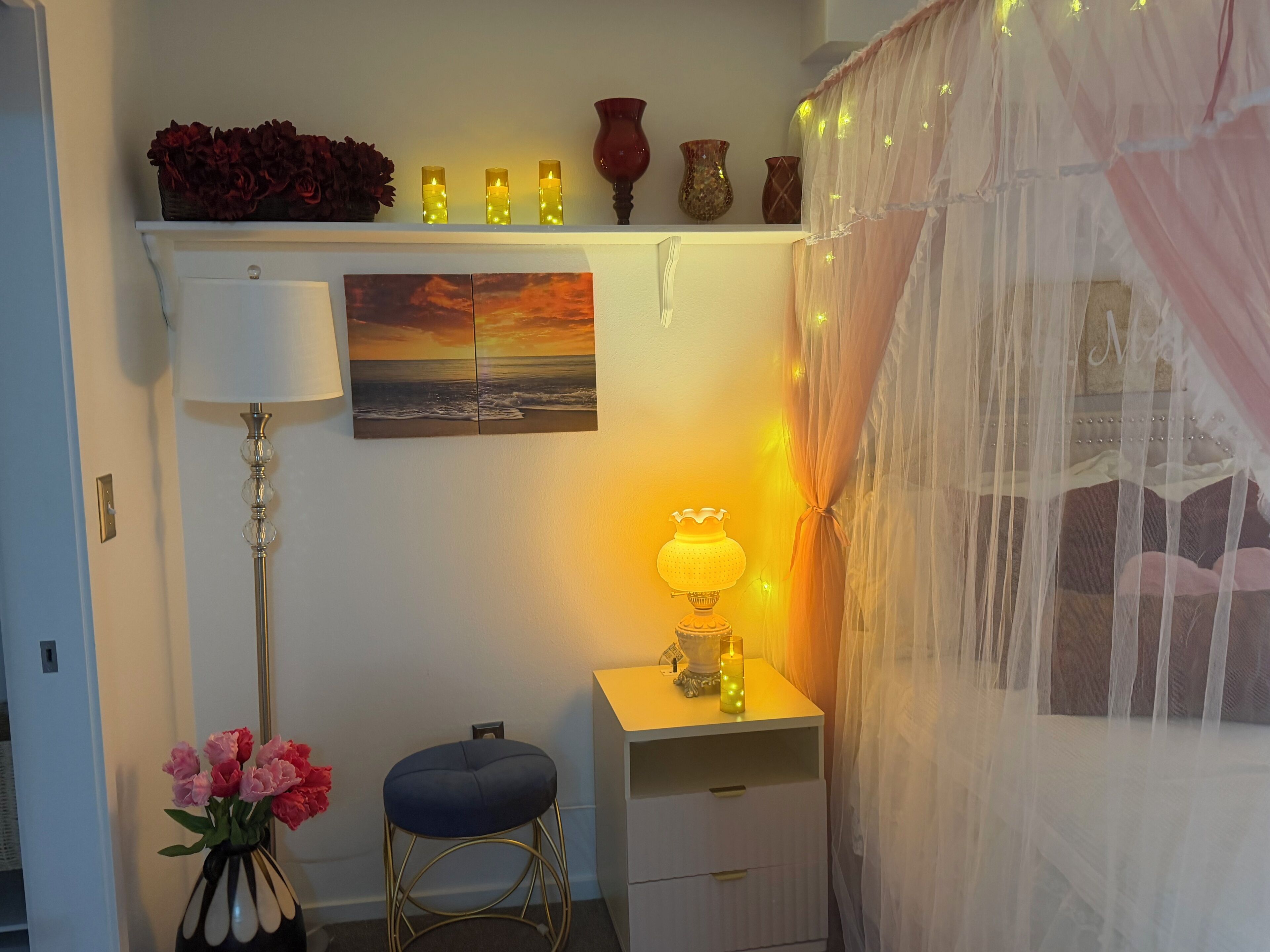 A room with a desk, a lamp, a vase with flowers, a painting, and a bed with a canopy.