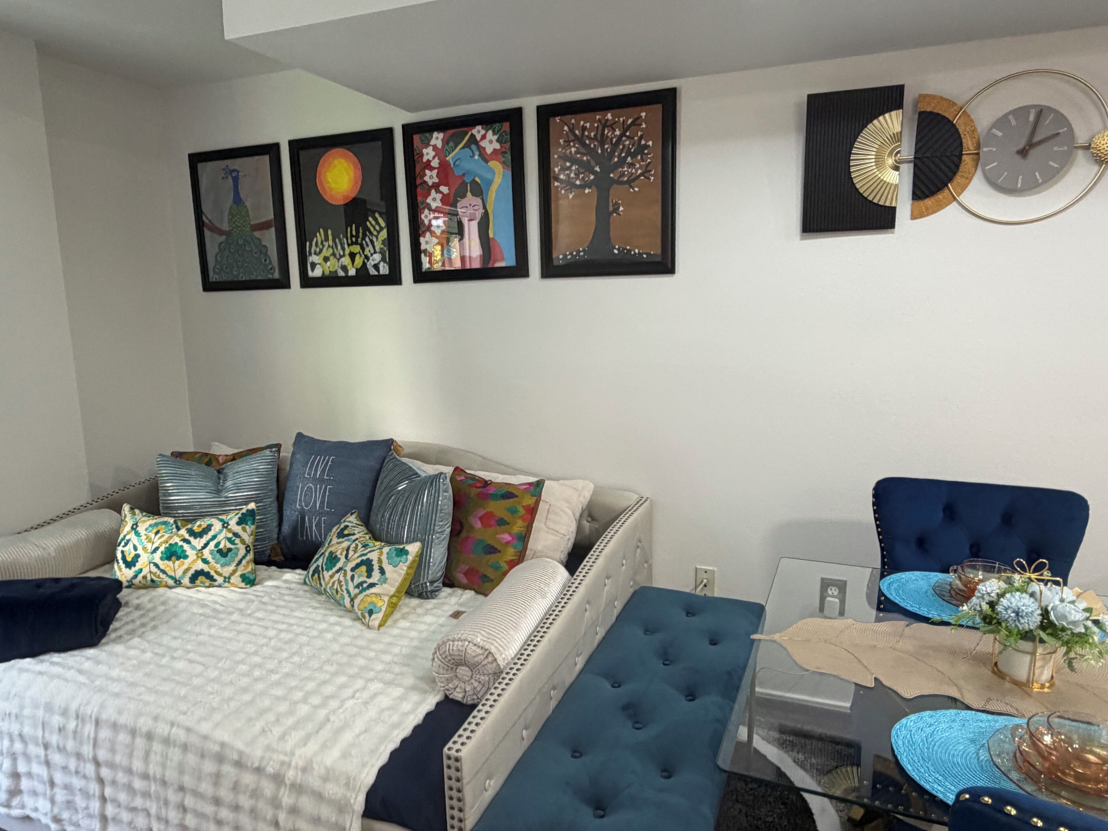 A bedroom with a bed, a blue armchair, a dining table with a vase, and wall art.