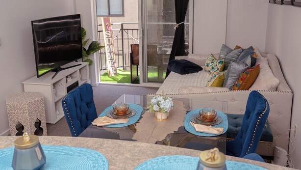 Un salon avec un canapé, une télévision, une table à manger avec des sets de table et un balcon orné d’une pancarte indiquant « SUMMER ».