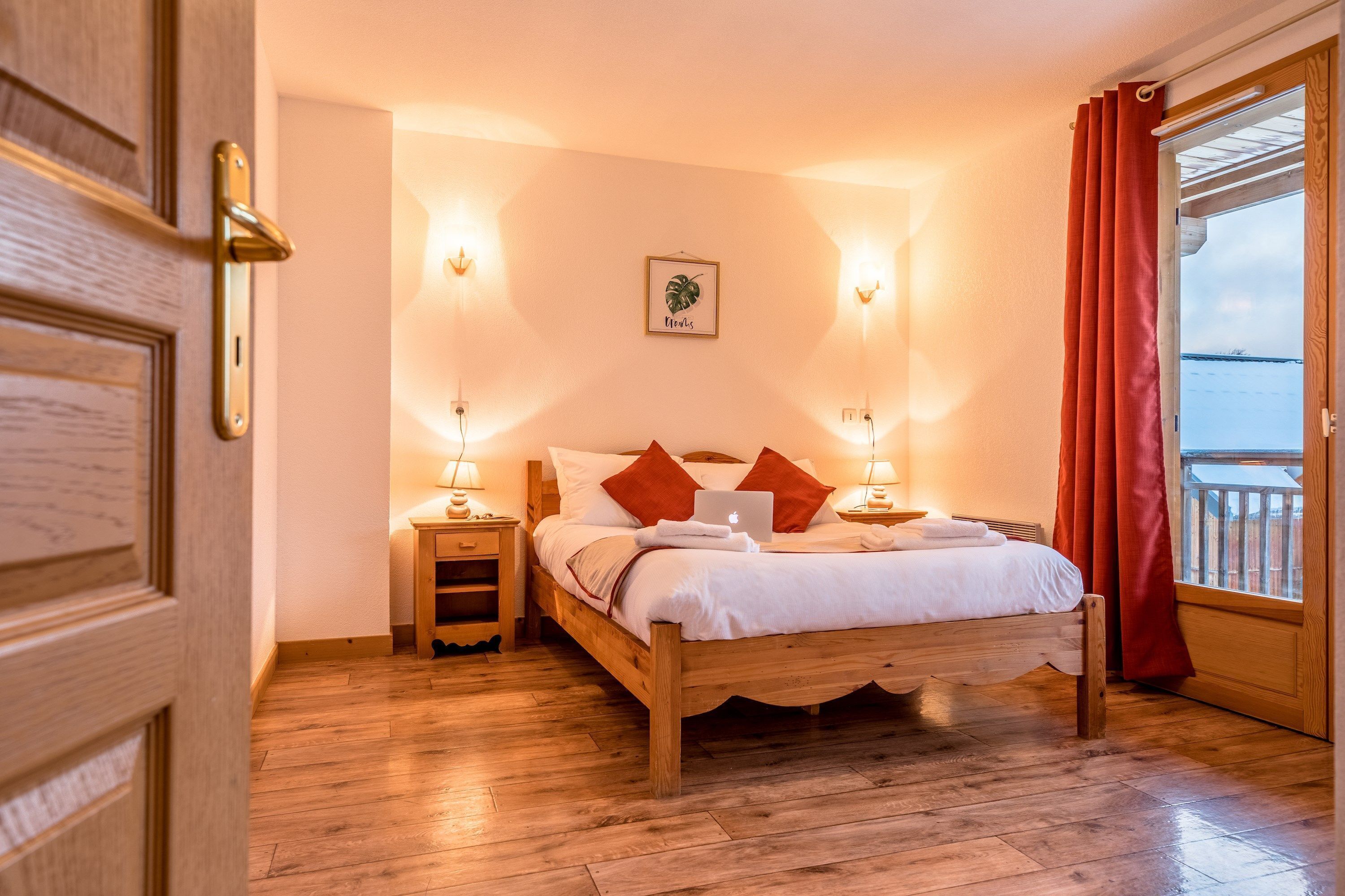 A wooden bed with white bedding, red pillows, and a laptop on the nightstand. There are two wall-mounted lamps, a framed picture on the wall, and a window with red curtains.