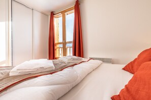 Ein ordentlich bezogenes Bett mit weißer und orangefarbener Bettwäsche, ein Fenster mit roten Vorhängen und Blick ins Freie.