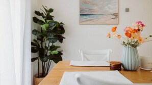 A dining table with a white tablecloth, a vase with flowers, a chair, a plant, and a painting on the wall. - West Grove Cottage Burnie (Parklands)