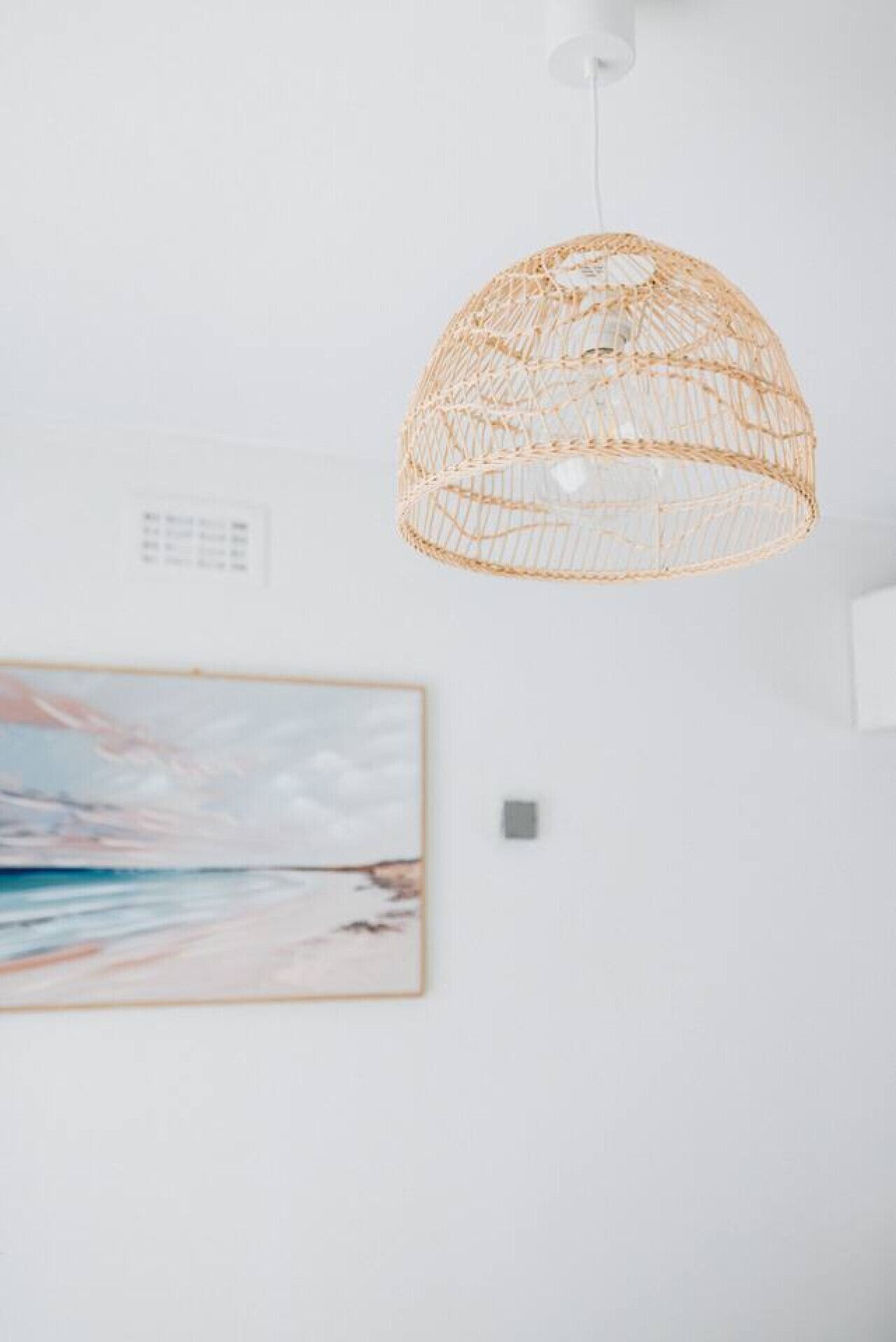 Una habitación con una lámpara colgante tejida, un cuadro enmarcado de una playa y una pared blanca.