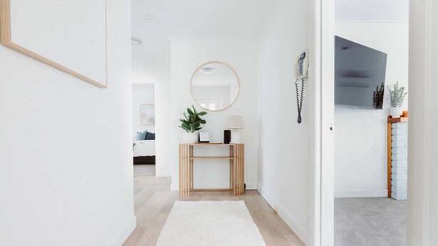 A hallway with a white rug, a wooden console table with a plant, a mirror, and a telephone. - West Grove Cottage Burnie (Parklands)