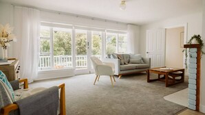 A spacious living room with a sofa, armchair, and coffee table, large windows with a view of greenery, and a white door. - West Grove Cottage Burnie (Parklands)