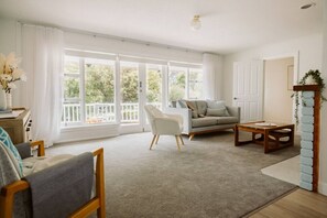 Ein großzügiges Wohnzimmer mit einer Couch, einem Sessel und einem Couchtisch, große Fenster mit Blick ins Grüne und eine weiße Tür.
