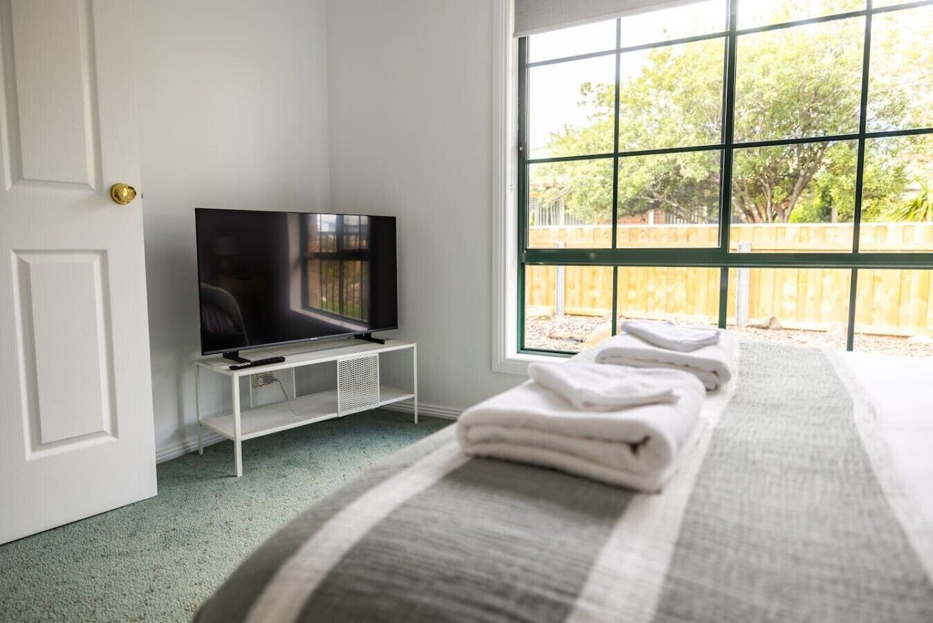 Une chambre d’hôtel avec un lit, un téléviseur posé sur un meuble et une fenêtre donnant sur des arbres et une clôture.