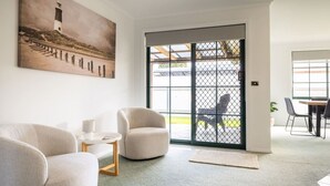 Ein Wohnzimmer mit Sofa, Sessel und einem kleinen Tisch mit Lampe. Ein großes Fenster bietet einen Ausblick nach draußen.