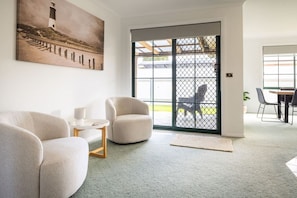 Ein Wohnzimmer mit Sofa, Sessel und einem kleinen Tisch mit Lampe. Ein großes Fenster bietet einen Ausblick nach draußen.