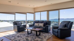 A modern living room with a sofa, two armchairs, a coffee table, and large windows offering a view of the sea.