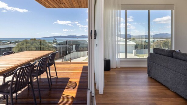 A modern living room with a wooden deck, large windows, and a view of the sea.