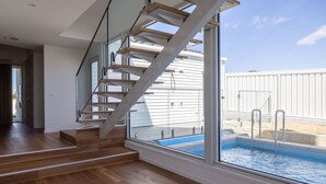 A modern interior with a wooden staircase, a large window overlooking a swimming pool, and a view of a white building.