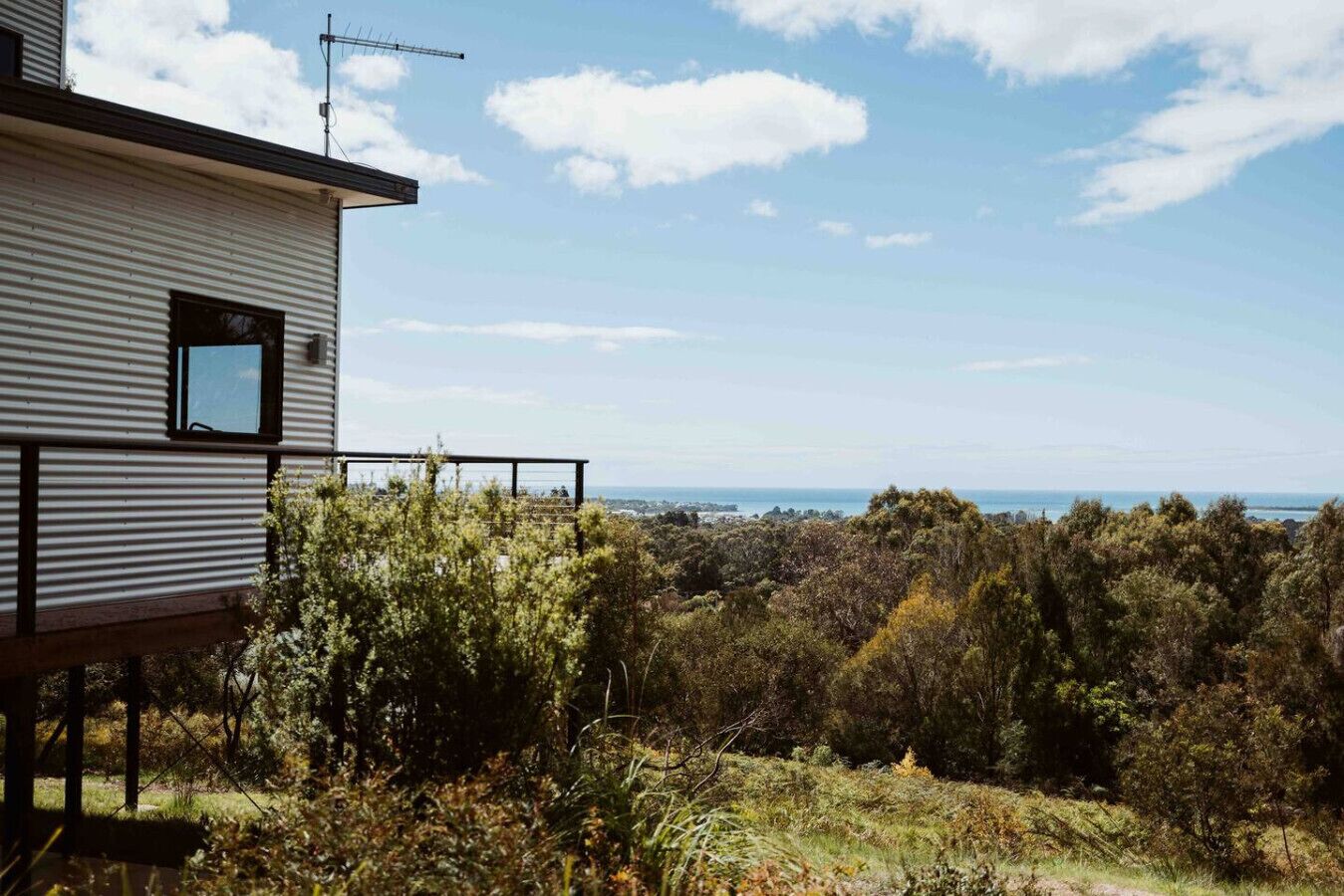 A modern building with a metal exterior, a small window, and a balcony overlooking a forested landscape with a distant view of the sea.