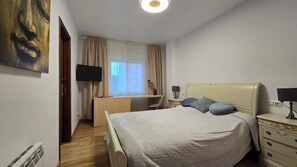 Makuuhuoneessa on sänky, yöpöydät, työpöytä, tuoli, televisio ja ikkuna, jossa on verhot.