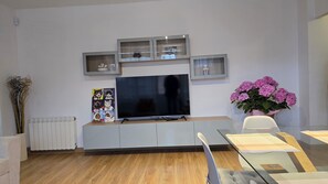 Moderni olohuone, jossa on litteänäyttöinen televisio, harmaat kaapit, patteri, maljakko täynnä kukkia sekä lasipöytä ja tuolit.