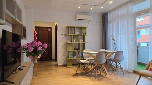 Ein modernes Wohnzimmer mit Fernseher, Holzboden, einem Essbereich mit Stühlen, einer grünen Regaleinheit und einem großen Fenster mit Vorhängen.