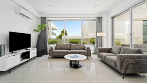 Ein modernes Wohnzimmer mit einem Flachbildfernseher, zwei Sofas, einem Couchtisch und einem großen Fenster mit Blick auf Grünflächen und Gebäude.