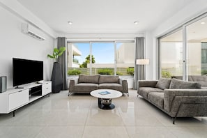 Ein modernes Wohnzimmer mit einem Flachbildfernseher, zwei Sofas, einem Couchtisch und einem großen Fenster mit Blick auf Grünflächen und Gebäude.