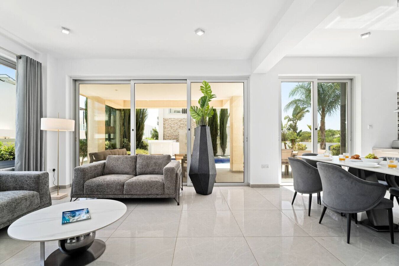 A modern living room with a grey sofa, a coffee table, and a dining area with chairs and a table set for breakfast.