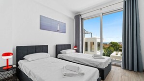 Ein Hotelzimmer mit zwei Betten, einem Nachttisch mit Lampe, einem Gemälde eines Leuchtturms und einem großen Fenster mit Blick auf Gebäude und Grünflächen.