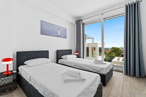 Ein Hotelzimmer mit zwei Betten, einem Nachttisch mit Lampe, einem Gemälde eines Leuchtturms und einem großen Fenster mit Blick auf Gebäude und Grünflächen.
