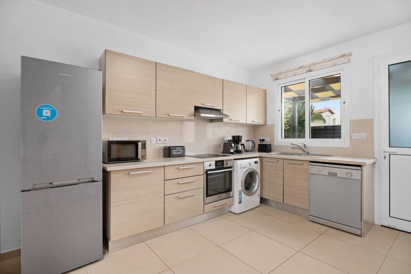 Een moderne keuken met een ingebouwde magnetron, oven en vaatwasser, lichte houten keukenkastjes en een raam met uitzicht.