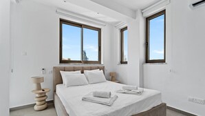Une chambre moderne avec un grand lit, deux fenêtres, une lampe de chevet et un thermostat fixé au mur.