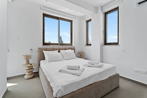 Ein modernes Schlafzimmer mit einem großen Bett, zwei Fenstern, einer Nachttischlampe und einem an der Wand montierten Thermostat.