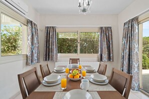 A dining room with a table set for six, surrounded by chairs, with a view of greenery outside through large windows. - Anthorina Gardens Retreat (Paralimni)