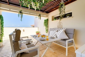 Eine Terrasse im Freien mit Tisch für Zwei, bequemen Sitzgelegenheiten, hängenden Pflanzen und Blick aufs Meer.