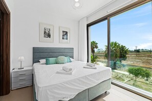 A modern bedroom with a large bed, a nightstand, a lamp, and a view of a landscape through a large window.