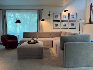 Ein Wohnzimmer mit einer Eckcouch, einem roten Sessel, einem Couchtisch mit Tablett, einer Stehlampe und gerahmten Bildern an der Wand.